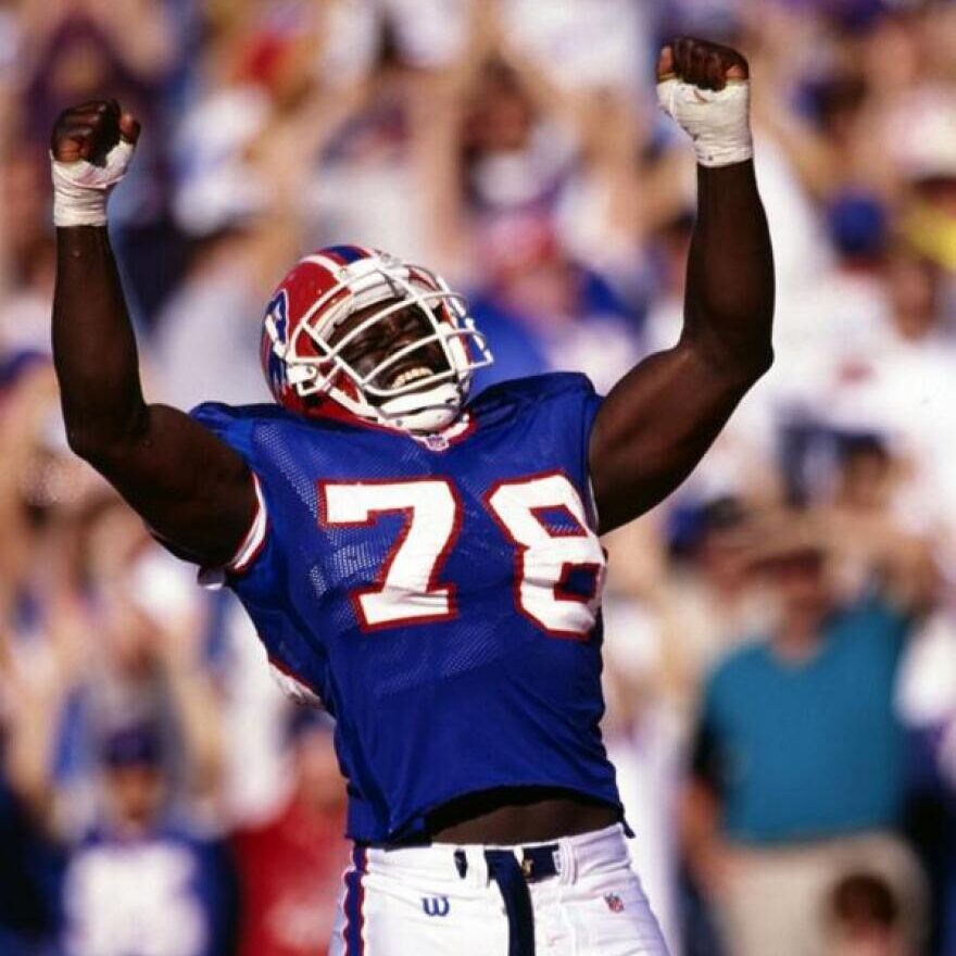 Football player in blue jersey with number 78 raises arms in celebration on the field, crowd blurred in background.